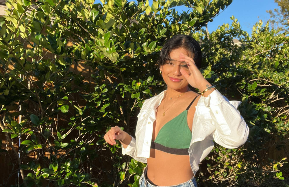 A woman wearing a white shirt, a green bralette, and jeans stands in front of a leafy bush with one hand raised to her face.