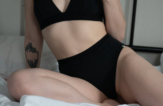 A tattooed woman wearing a black bralette and black underwear sits on a bed with white sheets.
