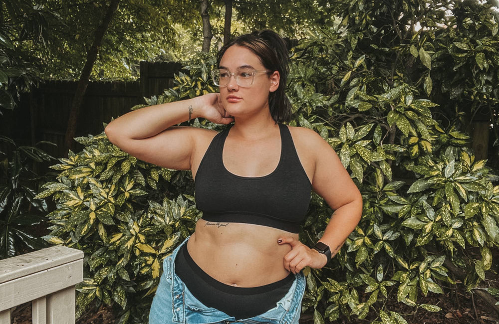 A woman with glasses wearing a black bralette, black underwear, and unbuttoned jeans stands in front of green shrubs