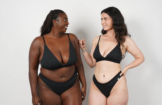 Two women wearing matching black bralettes and underwear sets stand in front of a white wall smiling at each other.