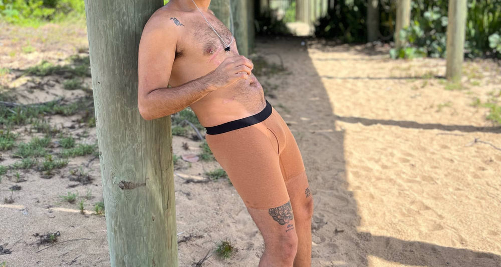 A man wearing a bucket hat, sunglasses, and beige boxer briefs leans against a wooden post on a beach.