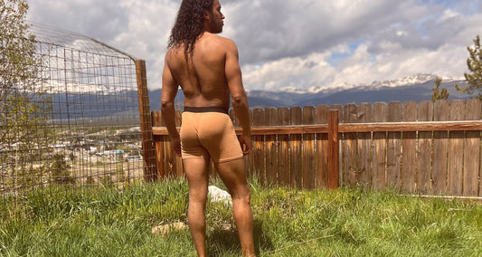  A curly-haired man wearing beige boxer briefs stands in a fenced backyard with a view of mountains and a cloudy sky. 