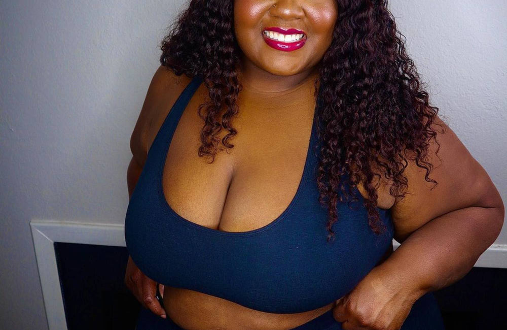 A smiling woman with long curly hair wearing a blue bralette and matching underwear stands with her hands on her hips.