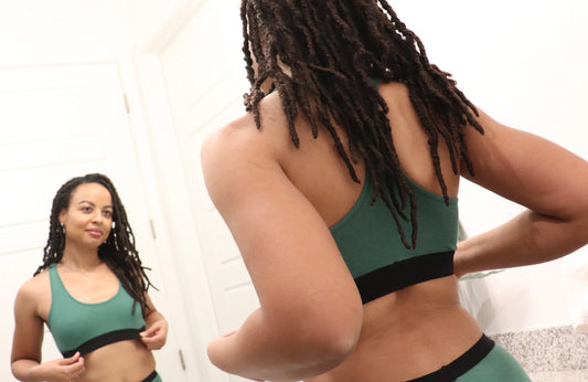A woman wearing a green bralette and matching underwear stands in front of a large bathroom mirror