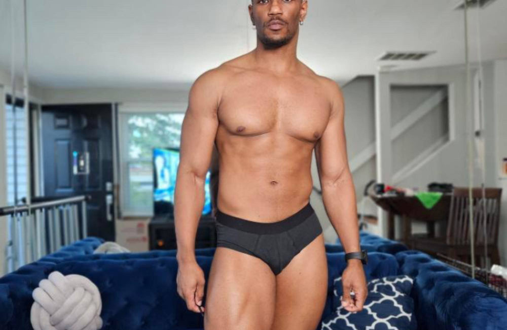 A man wearing black briefs stands in front of a blue couch and large wall mirror.