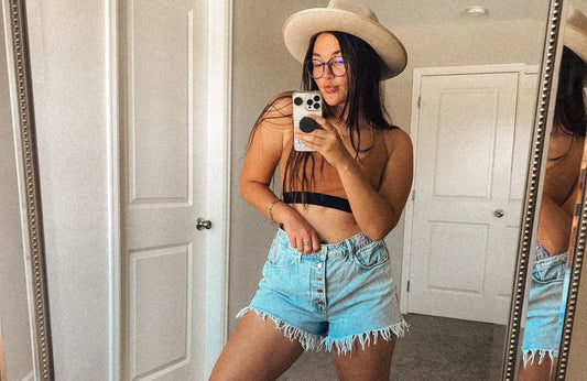 A woman with long brown hair wearing a brown bralette, jean shorts, and a white hat takes a mirror selfie