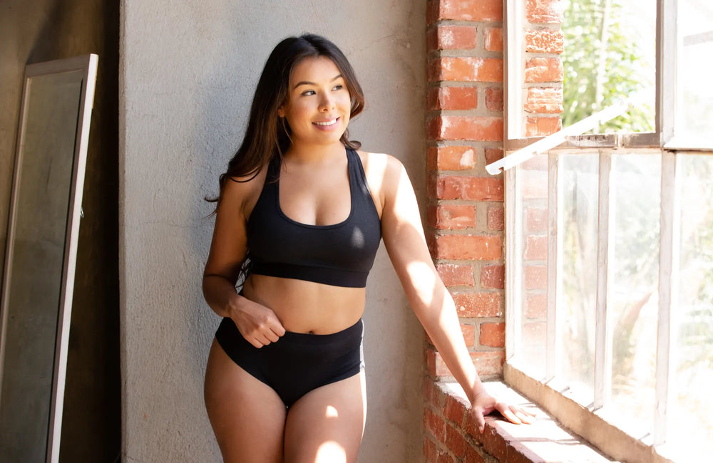 A woman with dark, long hair wearing a black bralette and black underwear stands next to a window and a brick wall.