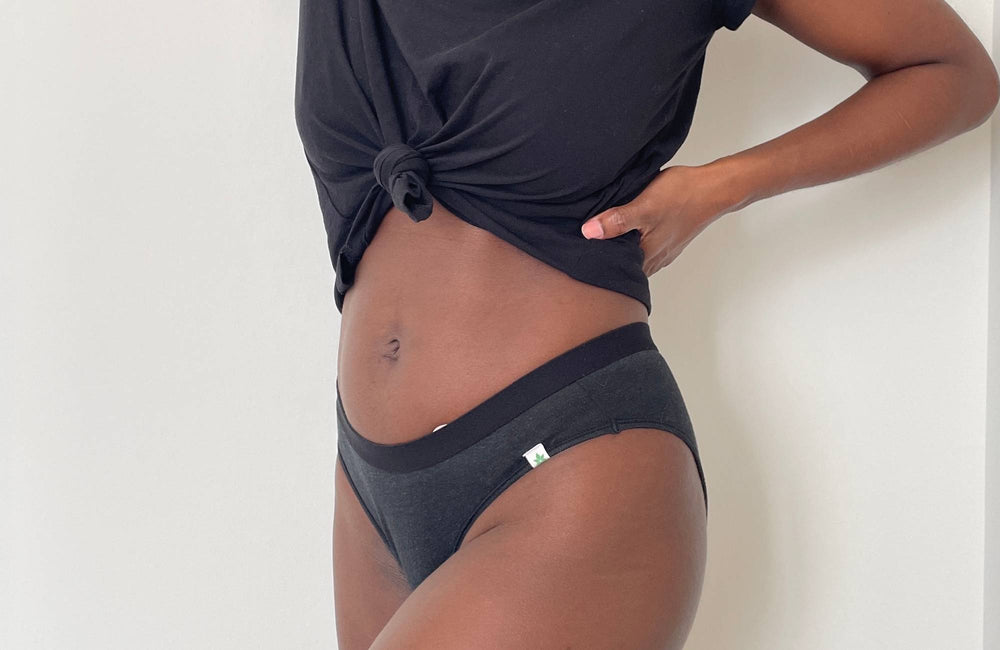 A woman wearing a tied black t-shirt and black panties poses with a hand on her waist in front of a white wall