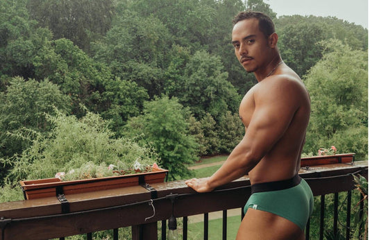 A man wearing green briefs stands with his hands on the railing of a balcony overlooking lush greenery.
