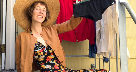 A woman wearing a floral print dress, a brown jacket, and a straw sun hat sits on a staircase holding up a pair of underwear on a hanger. 