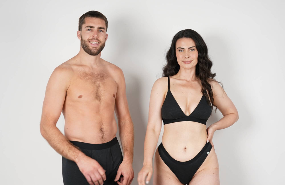 A man and a woman wearing black undergarments stand next to each other in front of a white background. 