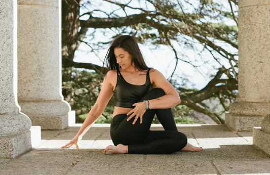 A woman wearing black yoga pants and a black bra stretches amidst stone columns in front of a large tree. 