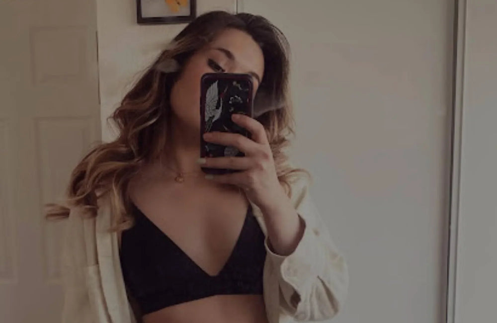 A woman wearing a black bralette and unbuttoned shirt takes a photo with a phone.