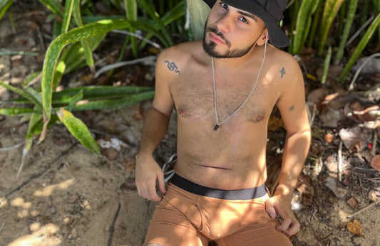 A man wearing a bucket hat and brown boxer briefs sits on the sand near a tree and plants