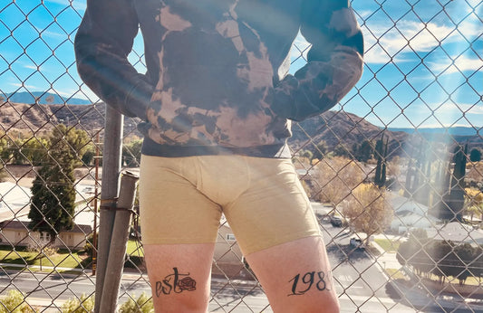 A man wearing yellow boxer briefs and a brown hoodie stands in front of a chain link fence