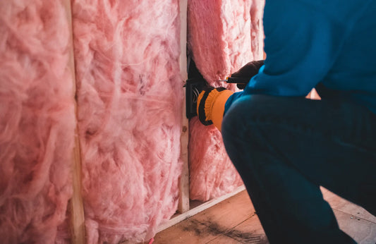  A person wearing a blue uniform and an orange glove holds a chunk of pink insulation in an unfinished wall.