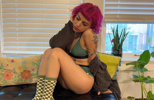 A woman with pink hair wearing a green bralette and matching underwear, a gray sweater, and checkered socks lounges on a leather sofa