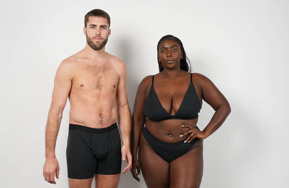  A man wearing black trunks underwear and a woman wearing a black bralette and matching underwear stand side by side in front of a white background