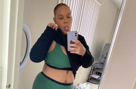  A woman pulls up her black sweater to expose her green bralette while she takes a mirror selfie with her tongue sticking out