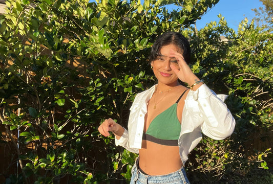 A dark-haired woman stands amongst bushes, wearing a green bralette, high-waisted jeans and an open white blouse.