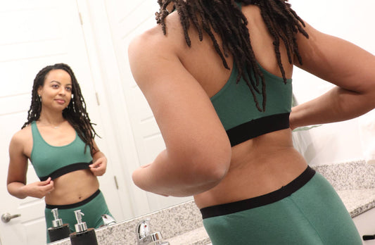 A woman with brown dreads wears green underwear and a green racerback bralette, while looking at herself in the mirror.