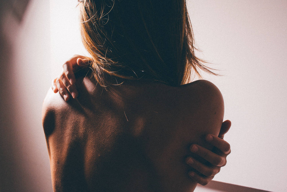 A topless woman faces away from the camera and wraps her arms around her back.