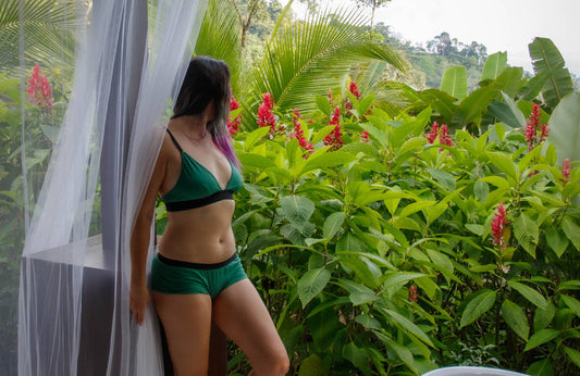 A woman wearing a green bralette and matching underwear stands outside near leafy green plants